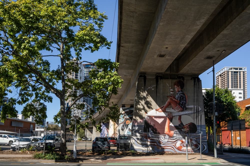 A large-scale mural on a concrete rail underpass pillar in Brisbane, framed by a street tree.